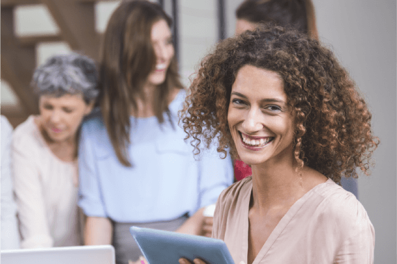 mujer que esta sonriendo sostiene una tablet y de fondo se ven tres mujeres que estan conversando entre ellas y estan viendo un computador