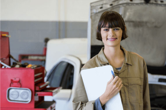 mujer con pelo corto sosteniendo una tabla y un laipz con un traje de mecanica y de fondo se ve una caja de herramientas y un auto blanco desarmado
